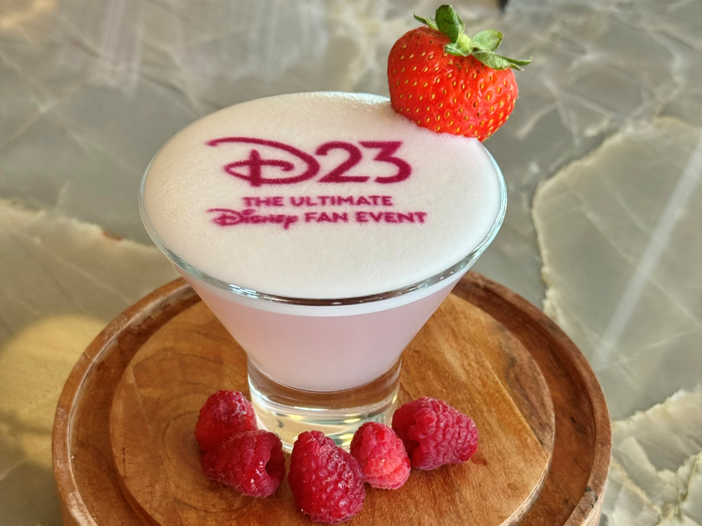 D23ロゴがフォームに印字された淡いピンクのカクテル、いちごとラズベリーを添えて