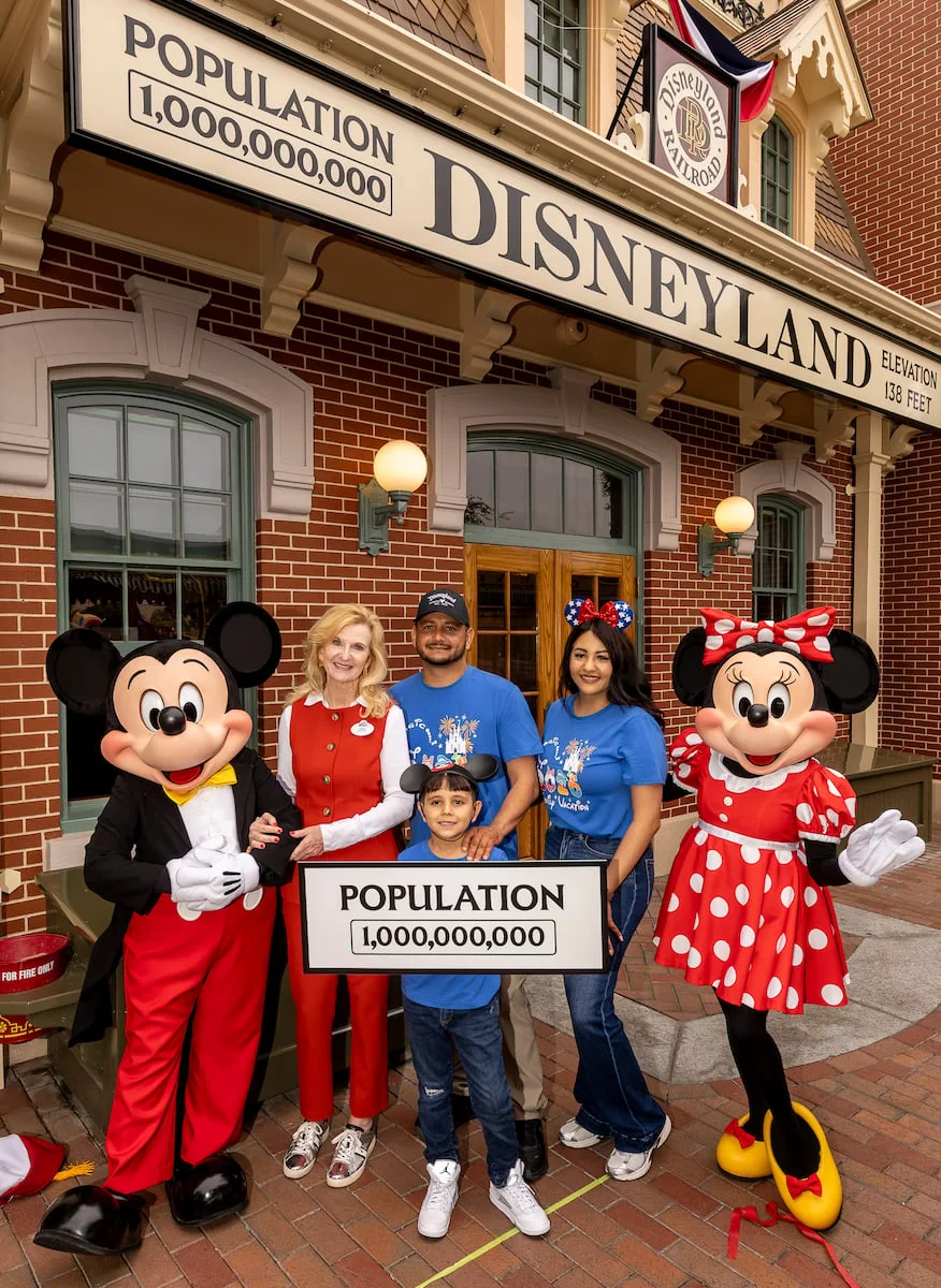 ディズニーランド・レイルロード駅にPOPULATION 1,000,000,000と更新されたサインの前でロブレス一家とミッキー・ミニーが記念撮影