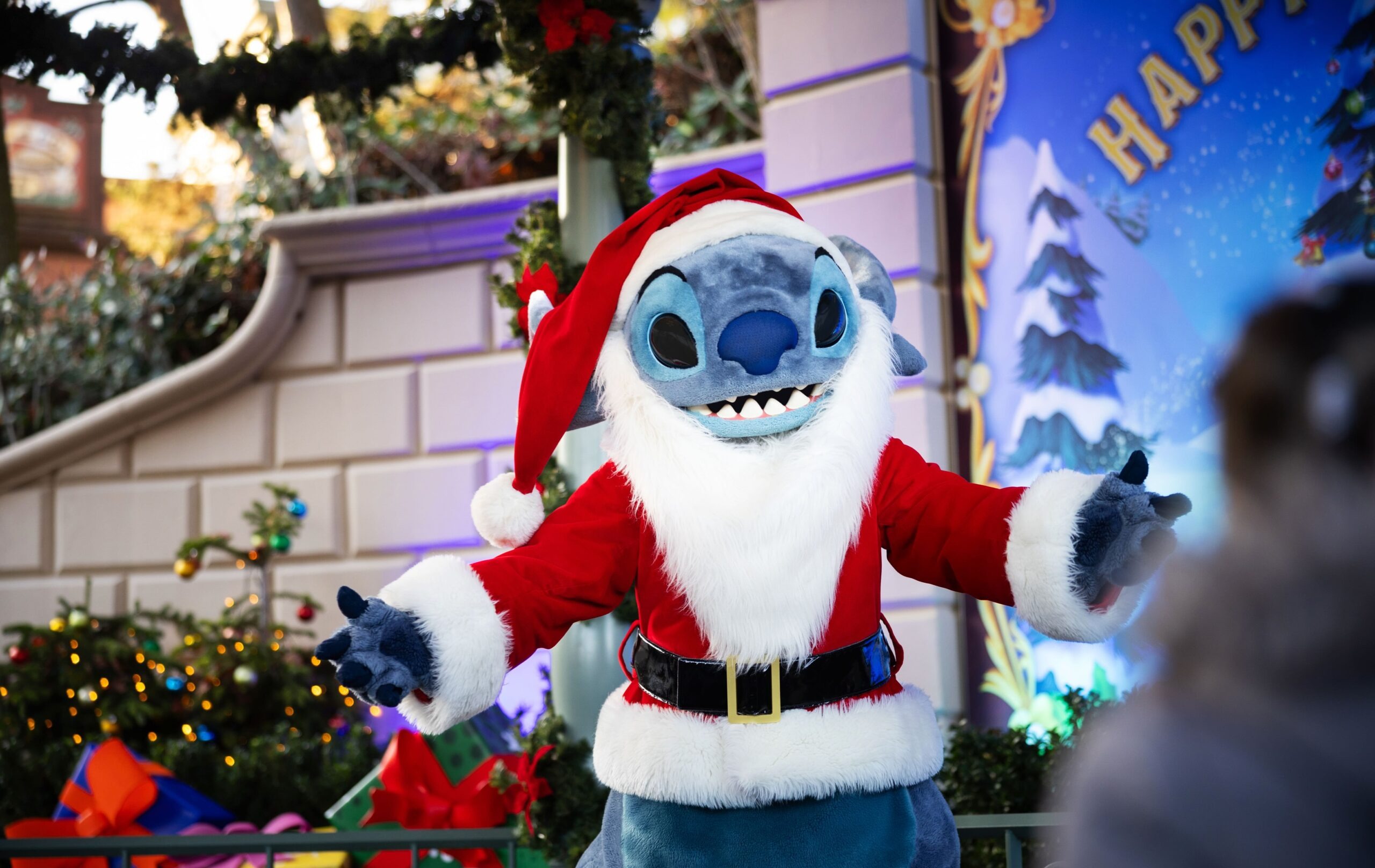 サンタクロースの衣装を着たスティッチの着ぐるみがクリスマス装飾の前でゲストを迎える様子